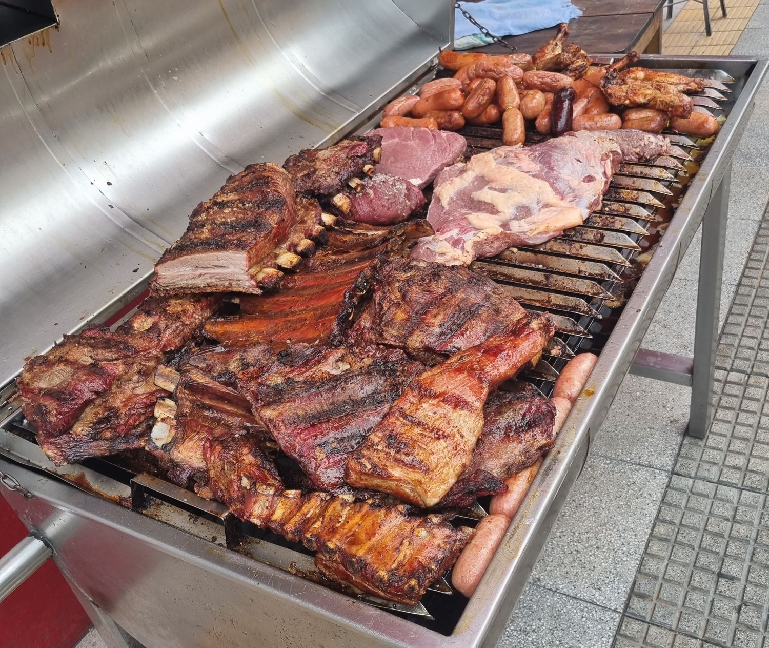 Großer Edelstahlgrill auf dem Gehsteig mit Rippen, Rindfleisch, Würsten und Hähnchen